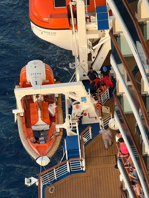 A lifeboat is stationed beside the Regal Princess cruise ship as several crew members in uniforms and red life vests prepare it, responding to an overboard incident. Passengers observe from the balcony while the scene unfolds over the ocean.