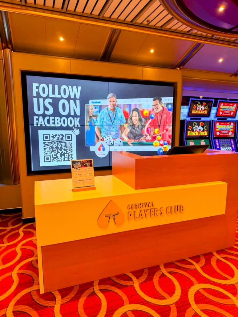 A Carnival Players Club desk stands on a red carpet. Behind it, a screen displays a “Follow Us on Facebook” message, a QR code, and a photo of people playing casino games—highlighting the excitement of cruise gamblers enjoying their luck. Slot machines are visible to the right, capturing the vibrant atmosphere found in cruise casinos.
