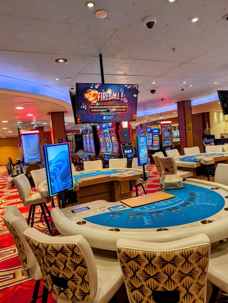 Casino interior with card tables, slot machines, and electronic screens. Chairs encircle the tables, and a digital sign overhead advertises a “Fireball” game. The carpet features a red geometric pattern, creating an inviting atmosphere for cruise gamblers eager to try their luck or experience the thrill of cashing out after a winning streak.