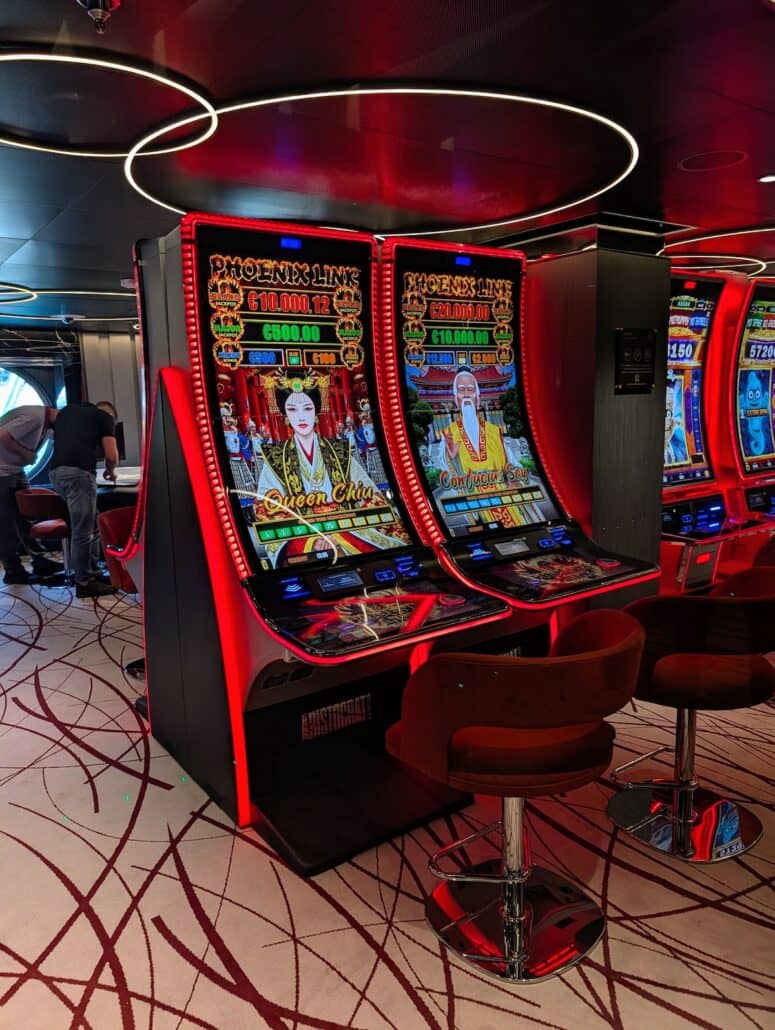 Two red-and-black slot machines with animated Asian-themed graphics and jackpot displays sit side by side in a casino. Red barstools are in front of the machines. Nearby, cruise gamblers are seen playing another slot machine, hoping for lucky spins and dreaming of cashing out big wins.