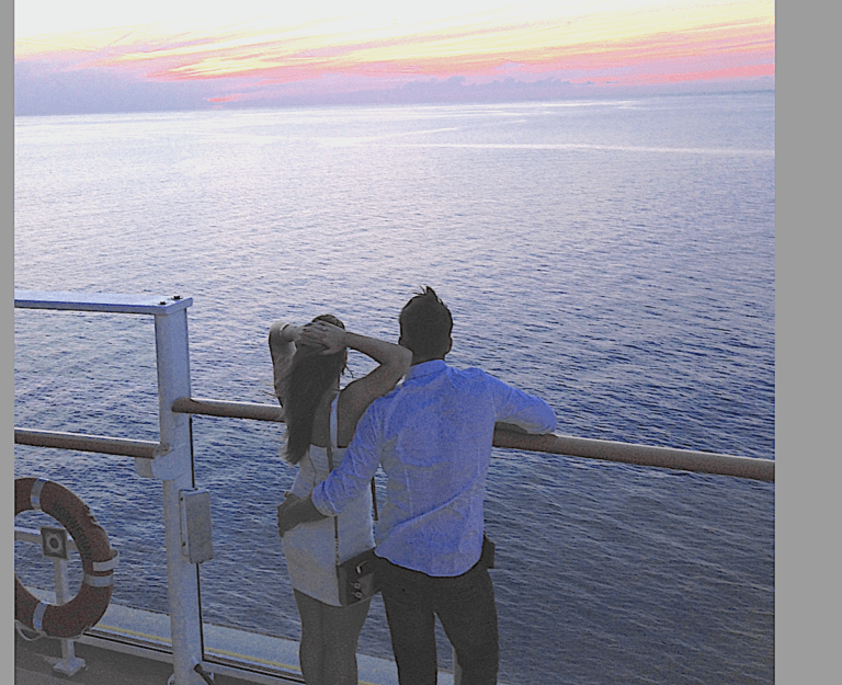A couple, perhaps first-time cruisers, stands together at the railing of a ship, looking out over the ocean at sunset. The sky is colorful with pink and orange hues, and a life preserver is visible on the left side of the image. Photo by Richard Simms