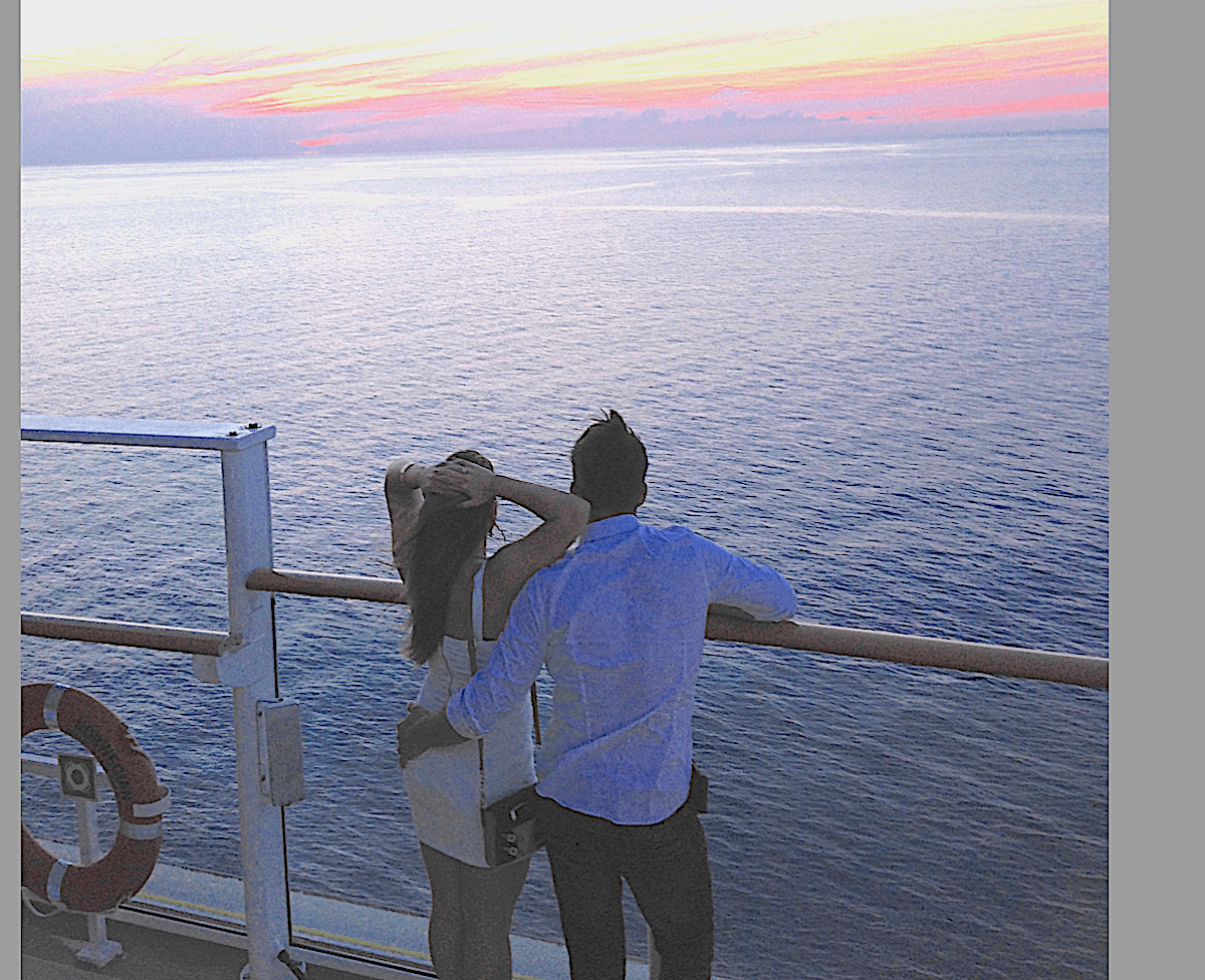 A couple, perhaps first-time cruisers, stands together at the railing of a ship, looking out over the ocean at sunset. The sky is colorful with pink and orange hues, and a life preserver is visible on the left side of the image. Photo by Richard Simms