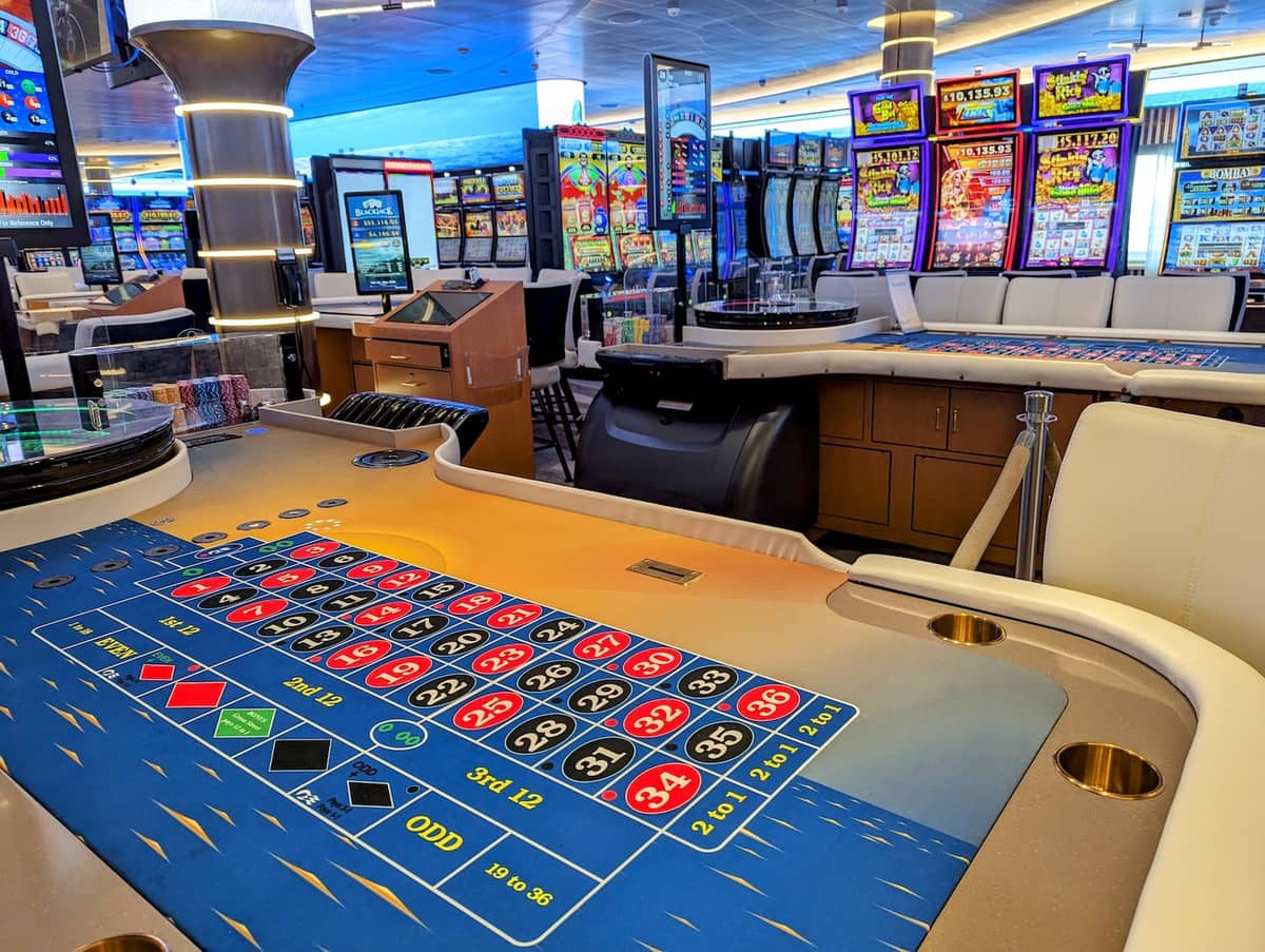 A brightly lit casino interior with a roulette table in the foreground and multiple slot machines with colorful screens in the background; white chairs are placed around the tables, creating an inviting atmosphere for cruise gamblers looking to try their luck before cashing out.