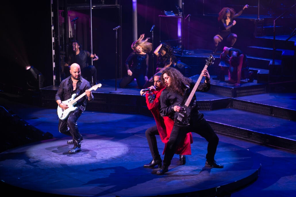 Three musicians perform energetically on stage with guitars and a microphone. Background dancers strike dynamic poses under purple lighting. Stage equipment is visible.