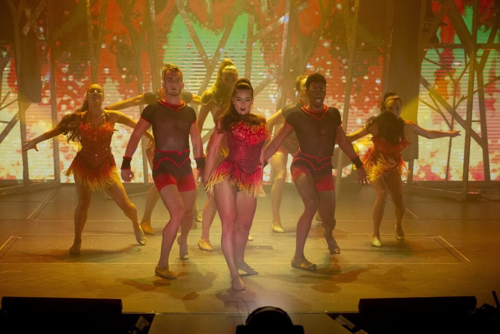 A group of dancers in coordinated red and yellow costumes perform on stage under warm lighting, with a vibrant, colorful background and dynamic poses.