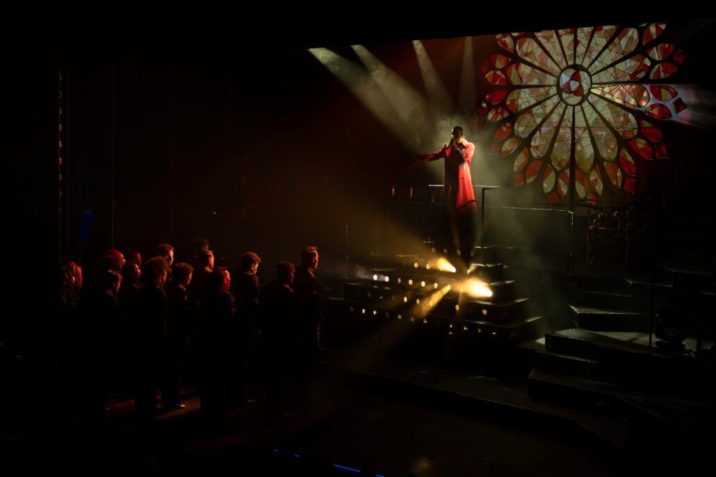 A performer in a red coat sings on a lit stage with a stained glass window backdrop, while a group of people stand in shadow, watching from the left.
