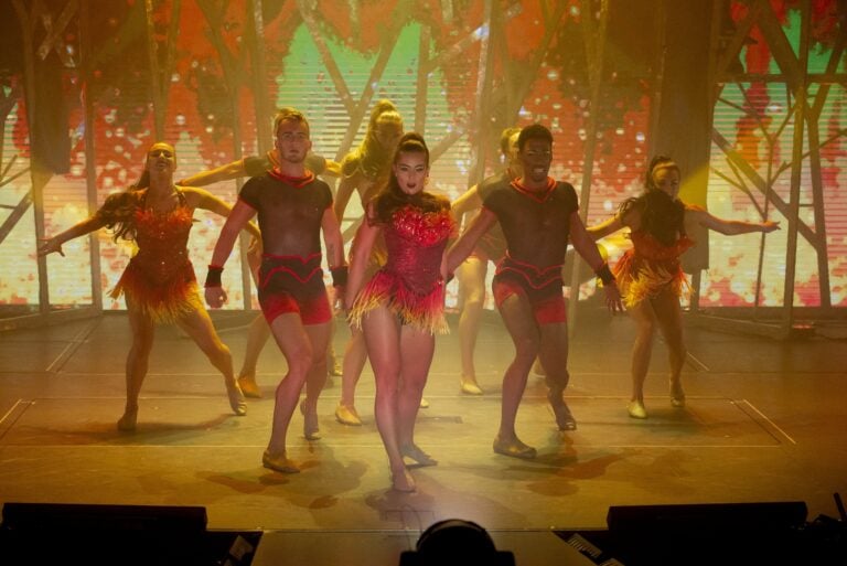 A group of dancers in coordinated red and yellow costumes perform on stage under warm lighting, with a vibrant, colorful background and dynamic poses.
