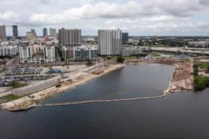 A city waterfront under construction with exposed soil, construction vehicles, and a barrier in the water; high-rise buildings and parking lots in the background under a cloudy sky.