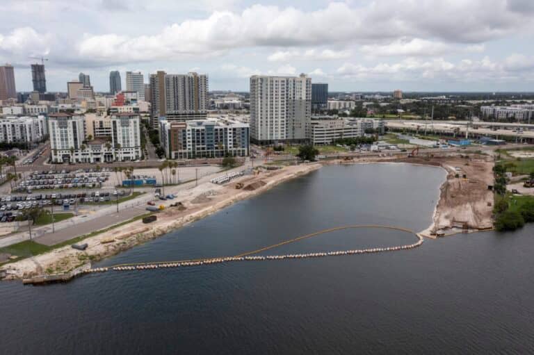 A city waterfront under construction with exposed soil, construction vehicles, and a barrier in the water; high-rise buildings and parking lots in the background under a cloudy sky.