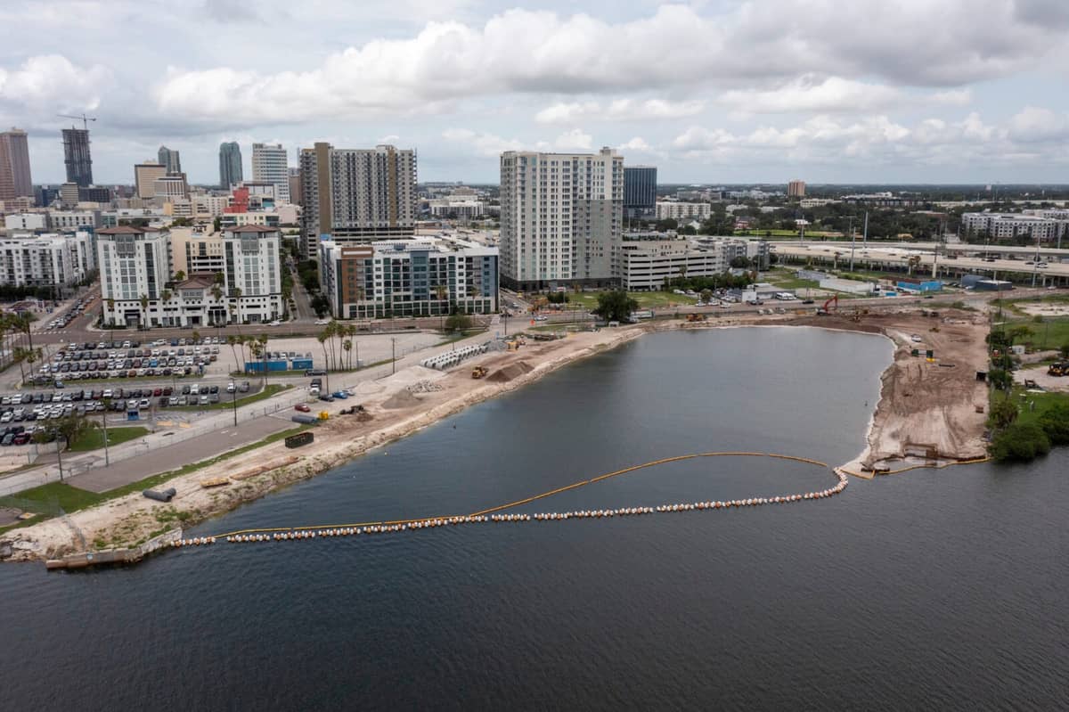 A city waterfront under construction with exposed soil, construction vehicles, and a barrier in the water; high-rise buildings and parking lots in the background under a cloudy sky.