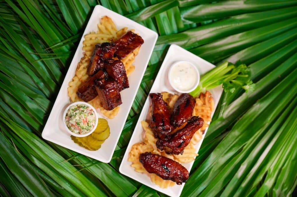 Two rectangular plates with barbecue glazed ribs or wings on waffle fries, served with coleslaw, pickle slices, celery sticks, and a side of ranch dressing. Plates are placed on large green palm leaves, adding a tropical flair reminiscent of Caribbean cliff jumps and the vibrant atmospheres found aboard Norwegian Cruise Line’s new waterpark adventures.