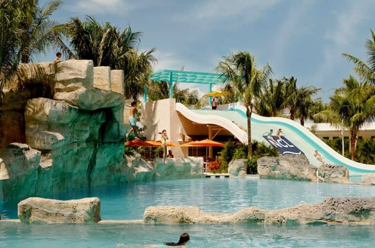 Children and adults enjoy a Caribbean waterpark with artificial rock formations, a pool, tall slides, and palm trees under a sunny sky. Some people are on slides while others swim or stand by the rocks, while thrill-seekers line up for exciting cliff jumps inspired by Norwegian Cruise Line’s signature adventures.