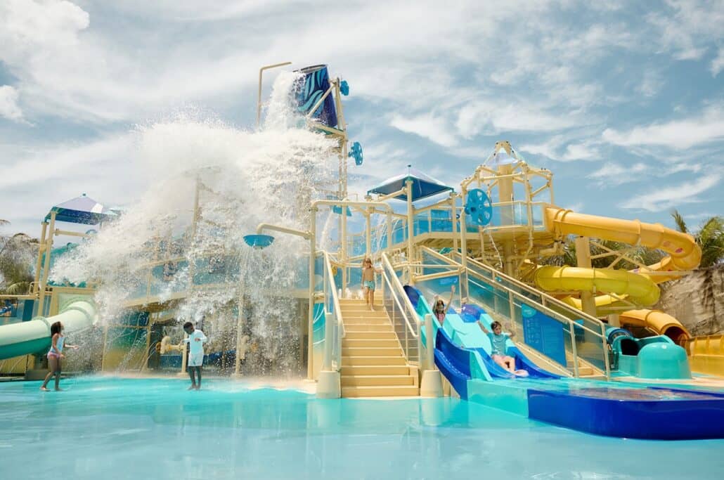 Children play at a colorful Caribbean waterpark with slides and climbing structures. A large bucket dumps water, splashing onto the area below. The sky is partly cloudy, and the park—featured on a Norwegian Cruise Line itinerary—is surrounded by tropical plants.