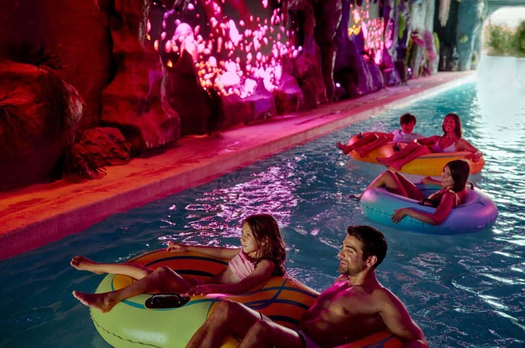 Four people ride inflatable tubes down a lazy river in an indoor Caribbean waterpark, passing by rock formations with glowing pink and orange lights on the wall. The atmosphere is relaxed and colorful—a perfect escape reminiscent of the vibrant experiences you’d find aboard a Norwegian Cruise Line adventure.
