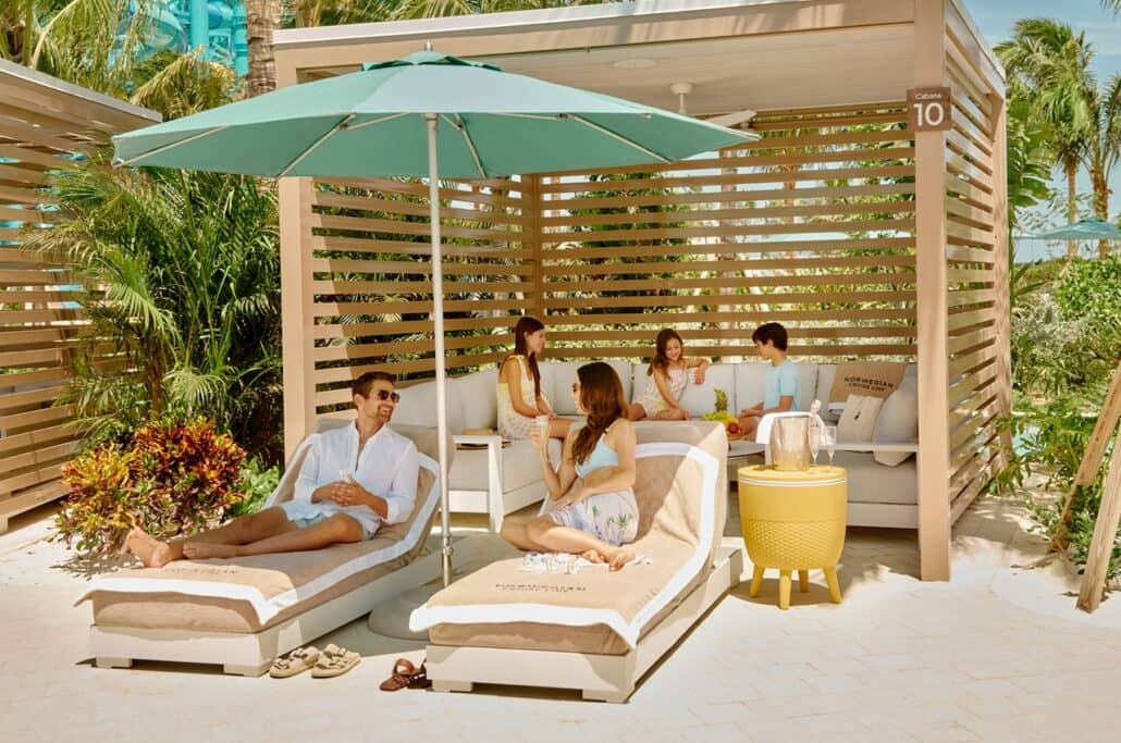 Four adults sit and relax on lounge chairs and a couch under a cabana at a tropical resort, reminiscent of the luxurious amenities found at a Caribbean waterpark. There is a large umbrella, green plants, and a small yellow table nearby. Everyone appears to be enjoying a sunny day.