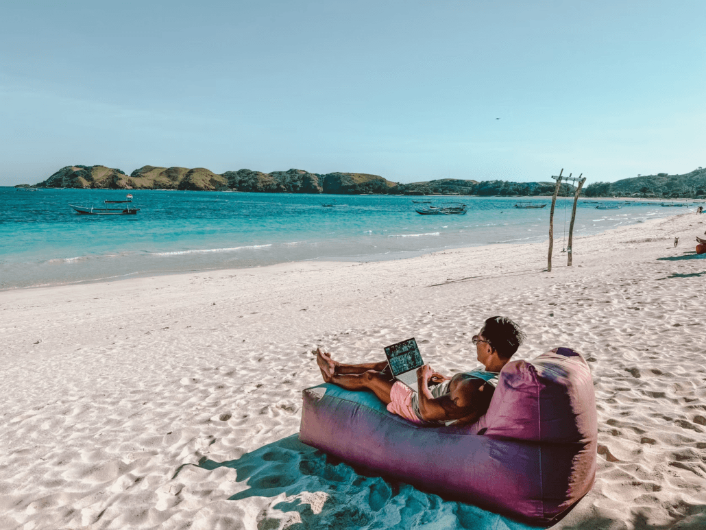A person sits on a pink bean bag on a sandy beach, using a laptop. The ocean and boats are visible in the background under a clear blue sky.