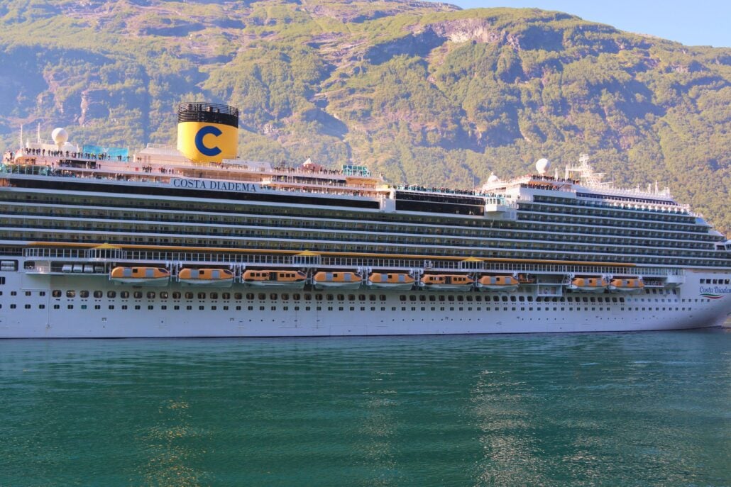 A large white cruise ship labeled “Costa Diadema” is docked near a lush, green mountainous shoreline, reflecting on calm, clear water under sunny conditions.