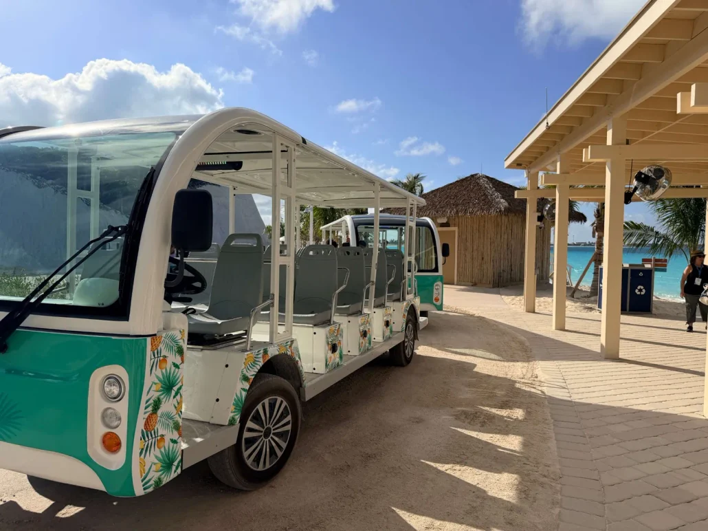 Open-air electric shuttle bus with tropical floral designs is parked on a sandy path near a wooden building and palm trees at RelaxAway in Half Moon Cay. The background features blue sky, scattered clouds, and turquoise water—inviting you to explore this beautiful island getaway with no extra costs for the free shuttle service.