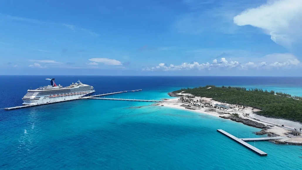 A large RelaxAway cruise ship is docked at a pier next to Half Moon Cay, a small island famed for its white sand beaches, clear turquoise water, lush green vegetation, and a few charming structures under a bright blue sky—the perfect spot to discover what’s free vs extra on your tropical getaway.