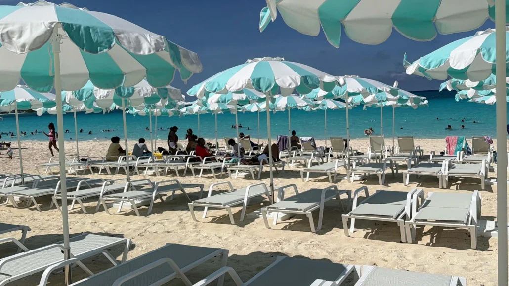 Rows of empty lounge chairs and turquoise-striped umbrellas line the sandy beach at Half Moon Cay. A group of people sits together near the water, enjoying some of the island’s many free activities. Swimmers and others are visible in the turquoise sea under a clear blue sky, all part of a perfect RelaxAway experience.