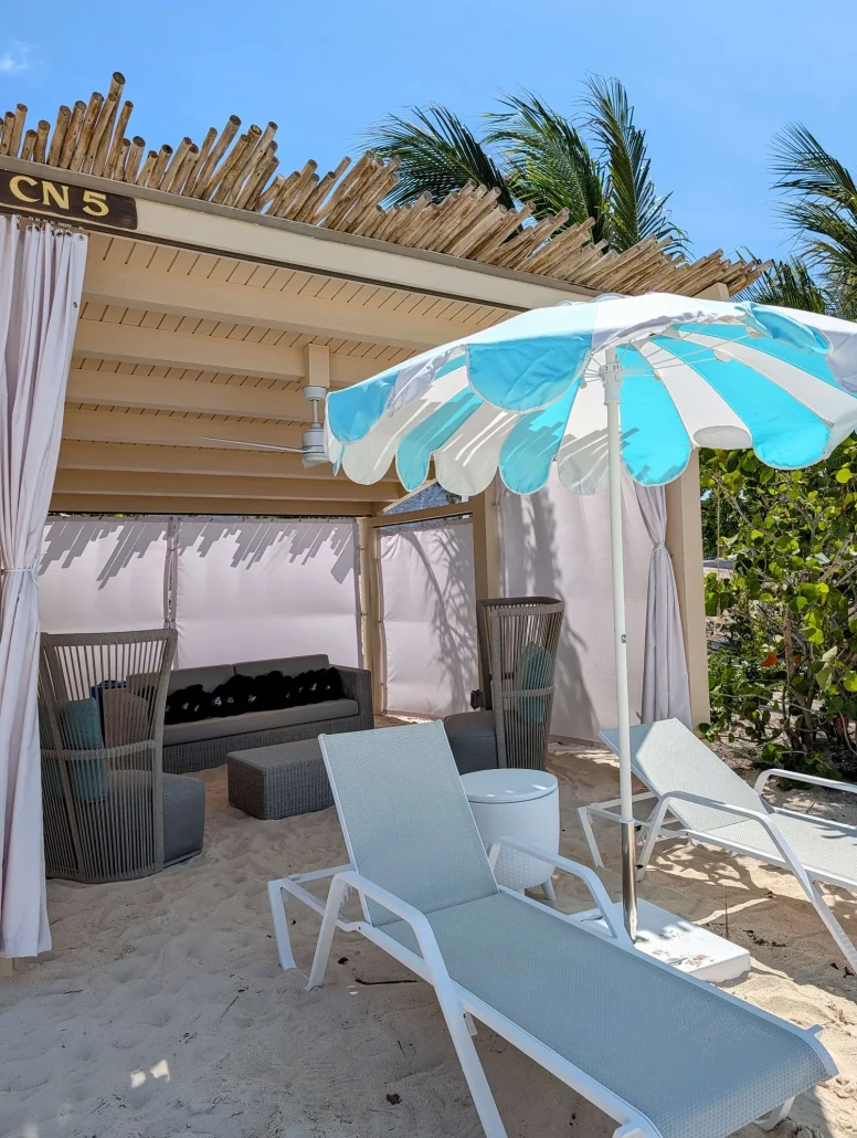 A RelaxAway beach cabana with lounge chairs, a shaded seating area, and a blue-and-white striped umbrella sits on sandy ground under a clear sky at Half Moon Cay, surrounded by palm trees and greenery—offering the perfect spot to experience the difference between free vs paid amenities in tropical comfort.