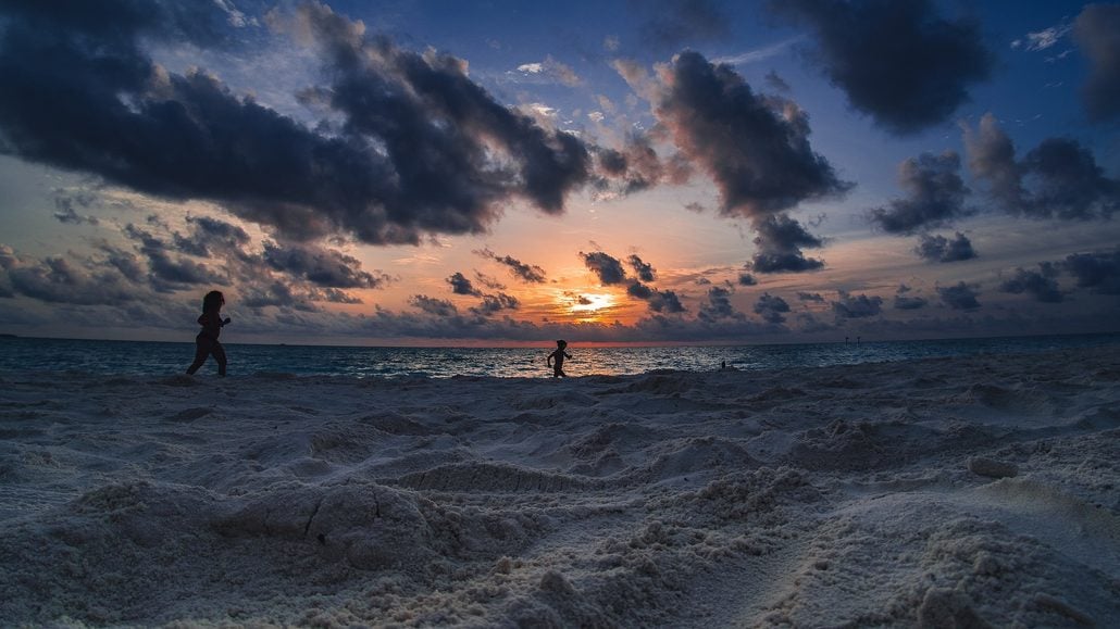 Two people walk along a sandy Seychelles beach at sunset, with dramatic clouds in the sky and the sun low on the horizon over the ocean. The sand is textured in the foreground.