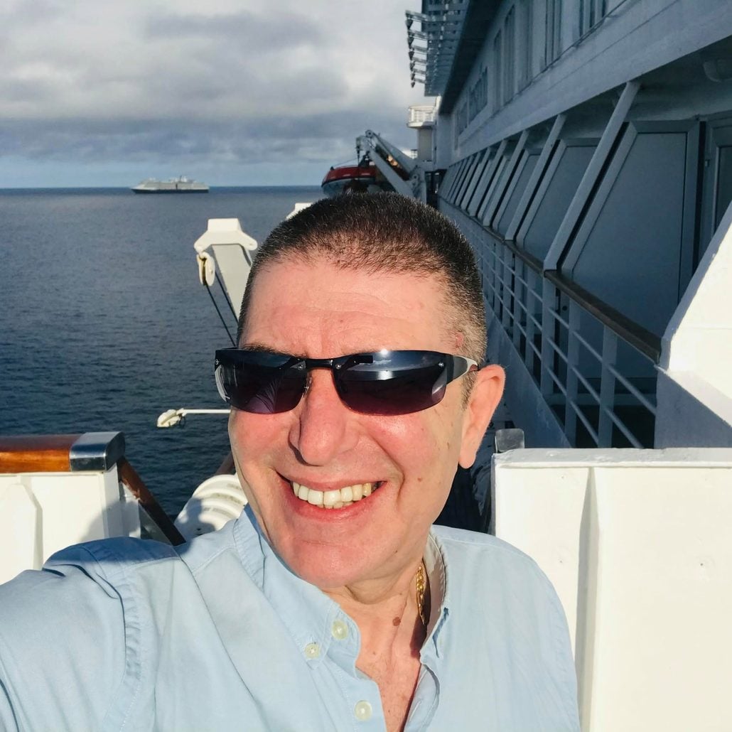 A man wearing sunglasses and a light blue shirt smiles for a selfie on the deck of a Carnival Cruise Line ship with calm sea and another ship visible in the distance under a cloudy sky.