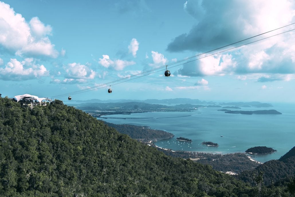 Cable cars travel above a lush green mountainside overlooking a blue sea with multiple islands, under a partly cloudy sky—a must-see highlight in any Langkawi travel guide for first-time visitors.