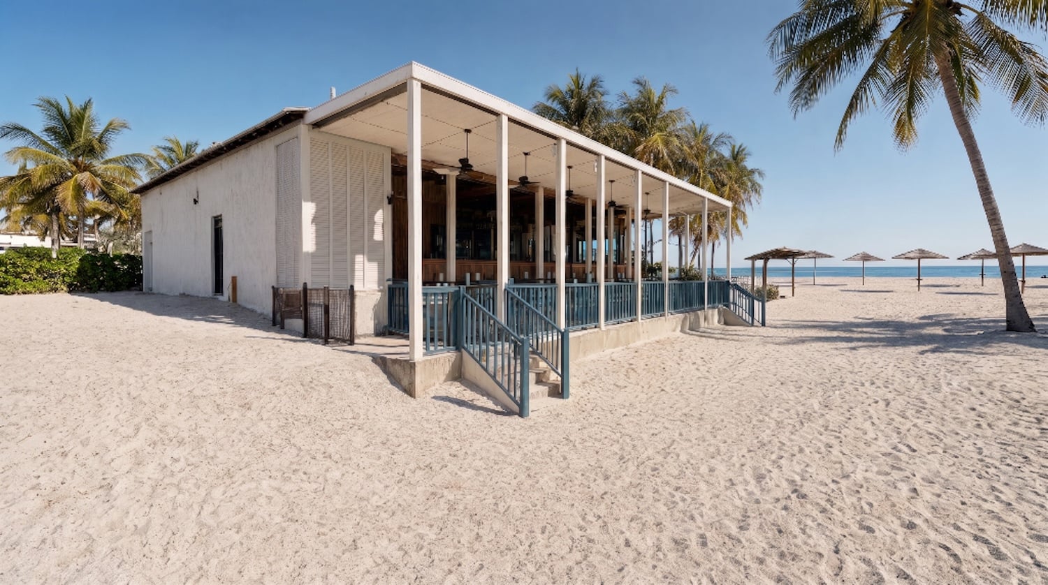 A beachside building with a covered patio stands on sandy ground, surrounded by palm trees. Several thatched sun umbrellas are set up on the beach near the ocean under a clear blue sky, creating the perfect tropical retreat at the Beach Club in Half Moon Cay, a beloved Holland America destination.