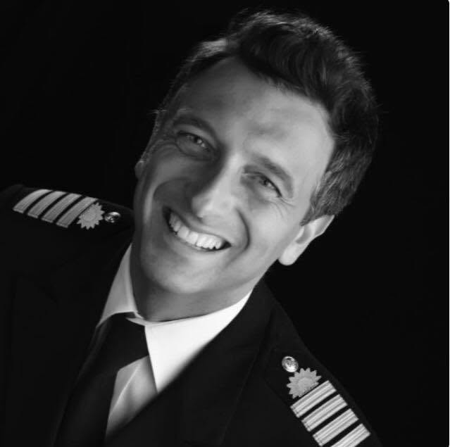 A man in a pilot uniform with epaulettes and a tie smiles at the camera against a black background. The image is black and white, featuring Captain Gennaro Arma of Princess Cruises, captured in his distinguished attire after 28 years of dedicated service.