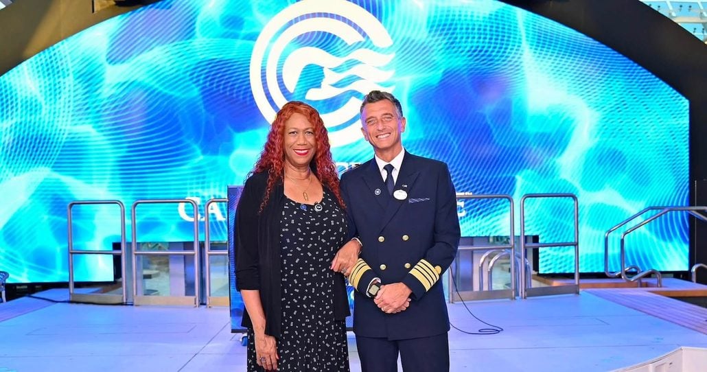 A woman in a black dress and a man in a Princess Cruises captain’s uniform—Captain Gennaro Arma—stand side by side, smiling, in front of a large blue digital backdrop with a wave logo, celebrating his 28 years with the company.