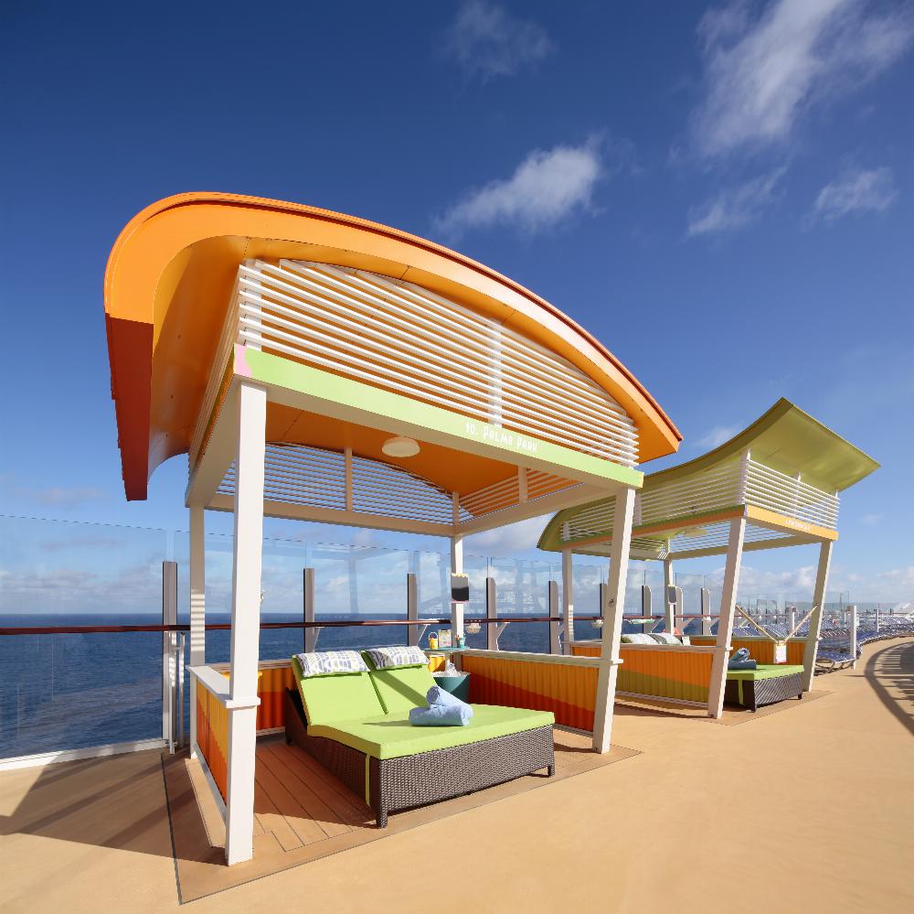 Two colorful cabanas with cushioned lounge beds sit on a deck overlooking the ocean under a clear blue sky at CocoCay. Glass barriers line the edge of the deck, and a distant shore is visible in the background, making this the ideal spot to place your bid for an unforgettable cabana experience.