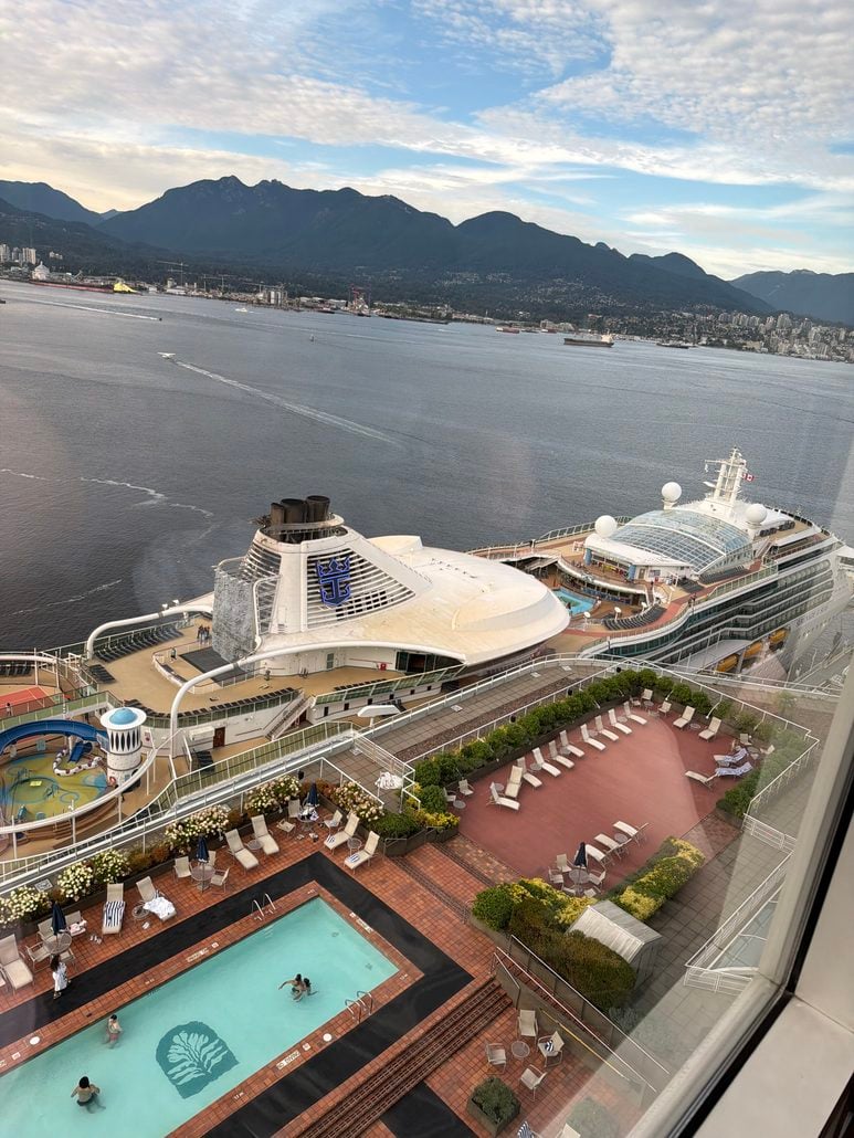 View from above showing a swimming pool with people, lounge chairs, and cruise ships docked at a port. Mountains and a city are visible across the water under a partly cloudy sky.
