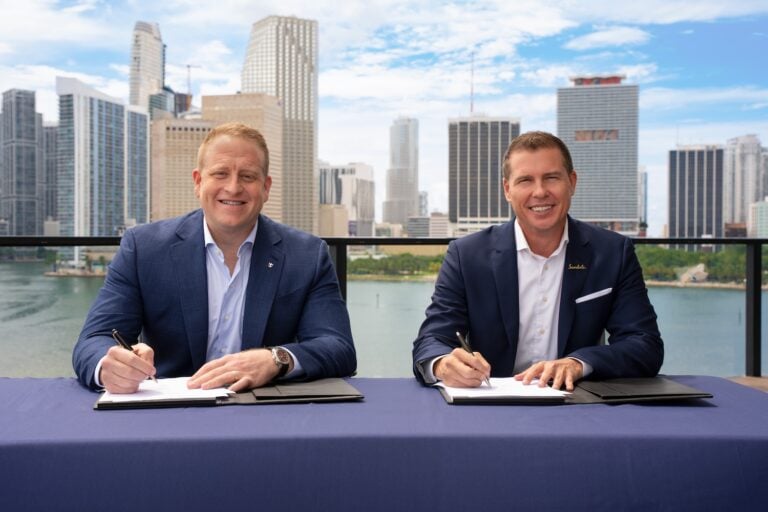 Two men in suits sit at a table, each holding a pen and signing documents. Tall city buildings and a waterfront are visible in the background on a cloudy day, evoking the elegance of a Royal Caribbean partnership event far from an all-inclusive resort setting.