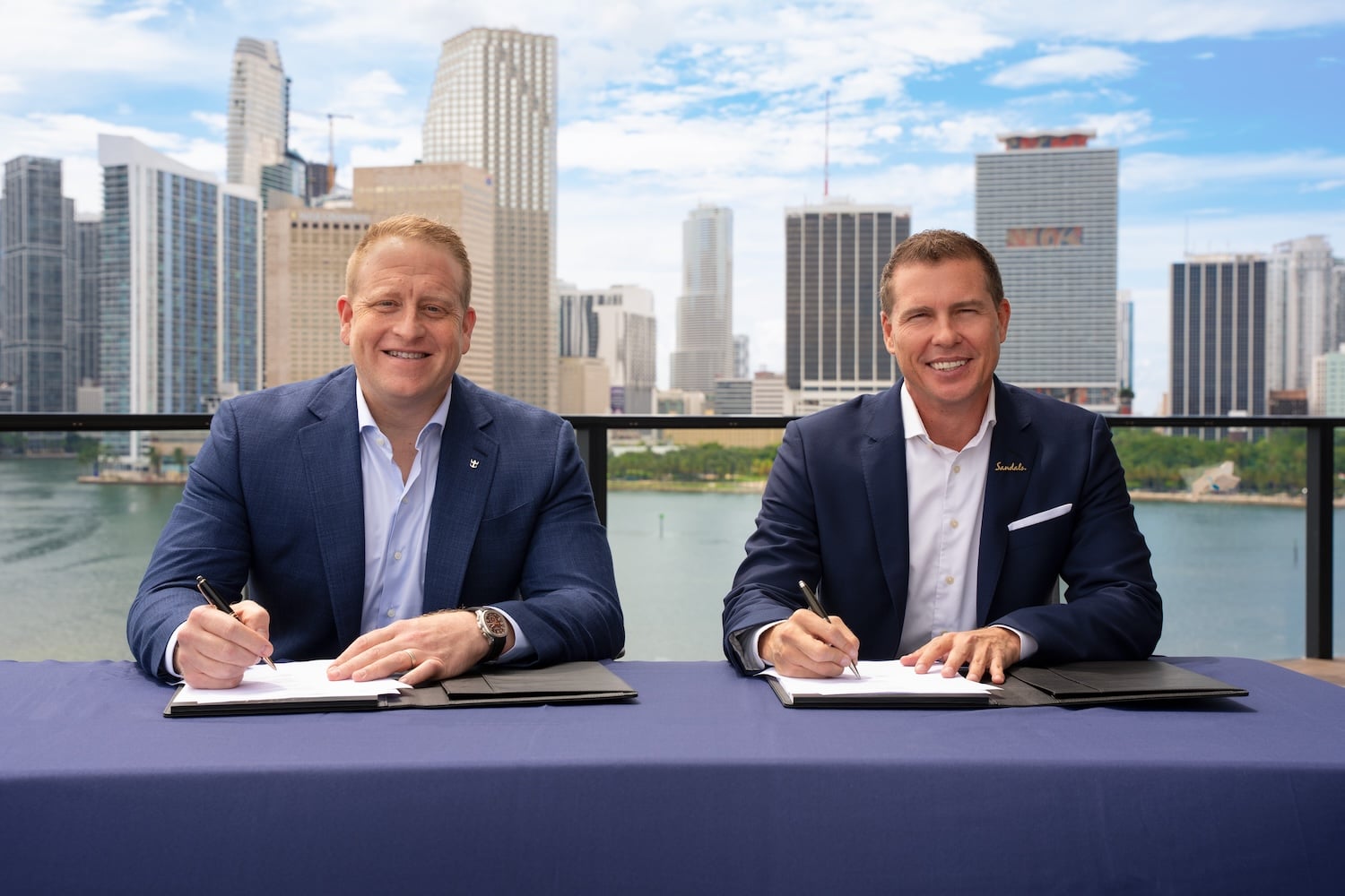 Two men in suits sit at a table, each holding a pen and signing documents. Tall city buildings and a waterfront are visible in the background on a cloudy day, evoking the elegance of a Royal Caribbean partnership event far from an all-inclusive resort setting.