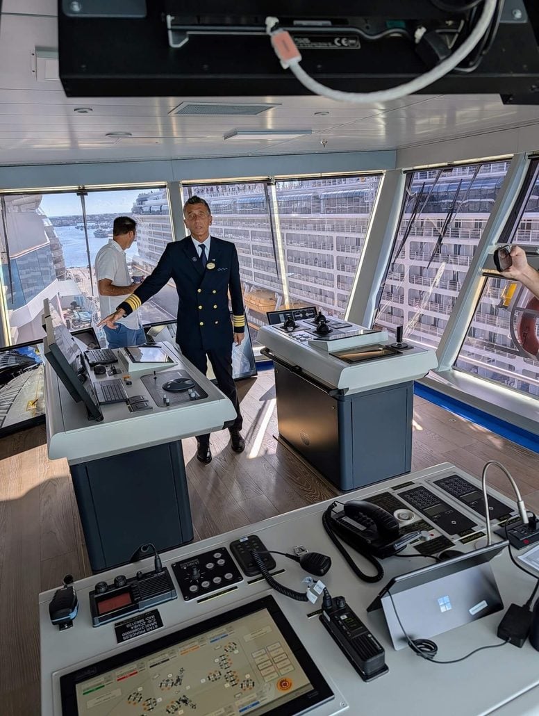 A uniformed officer stands on the bridge of a Princess Cruises ship near control panels. Captain Gennaro Arma, with 28 years of experience, is present alongside another individual, while someone in the foreground takes a photo. Large windows reveal the exterior and another docked cruise ship.