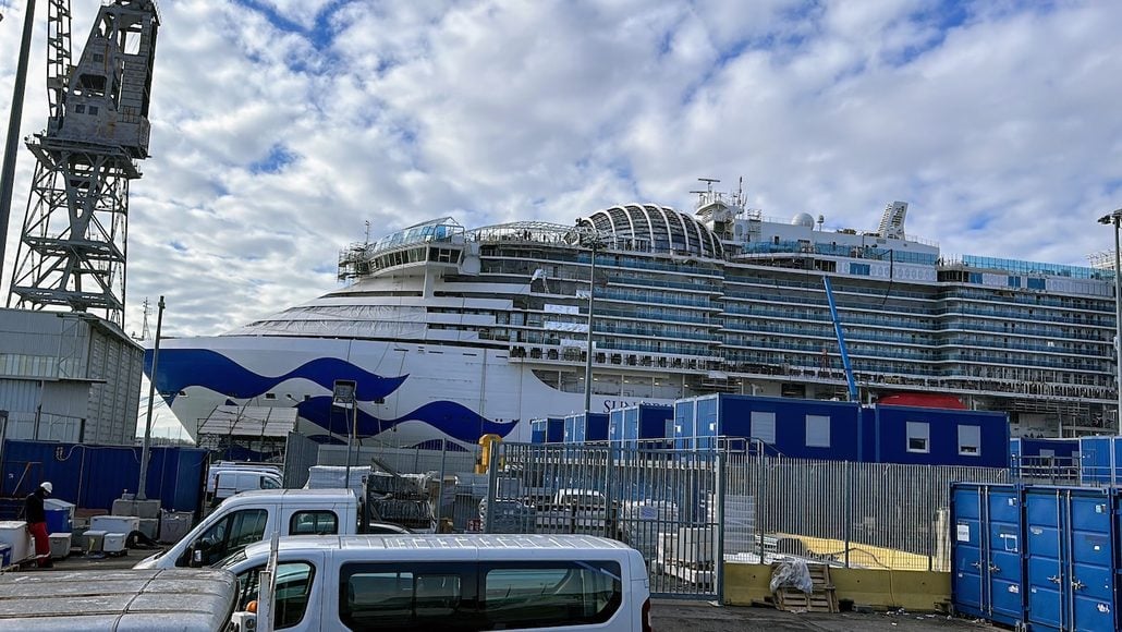 A large Princess Cruises ship, renowned for its 28 years of exceptional voyages under captains such as Captain Gennaro Arma, is docked at a port with blue portable buildings, parked vehicles, fencing, and a crane visible in the foreground under a partly cloudy sky.