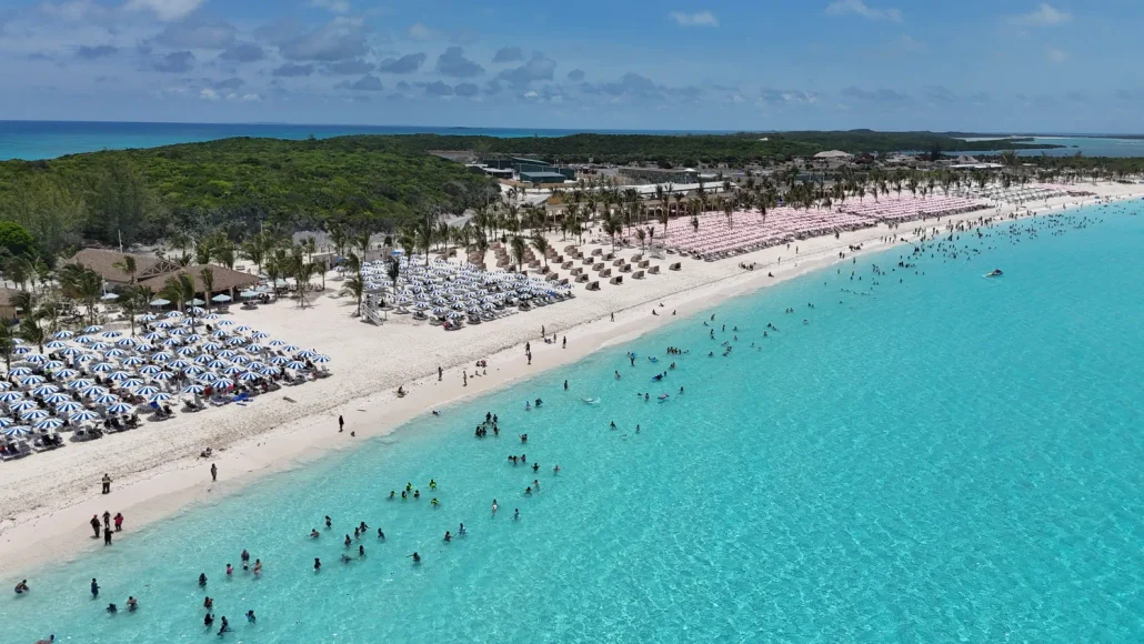 Aerial view of Half Moon Cay’s tropical beach with turquoise water, white sand, rows of lounge chairs and umbrellas—both free vs paid options available—people swimming, and lush green vegetation in the background under a blue sky with clouds, offering the perfect setting to RelaxAway.