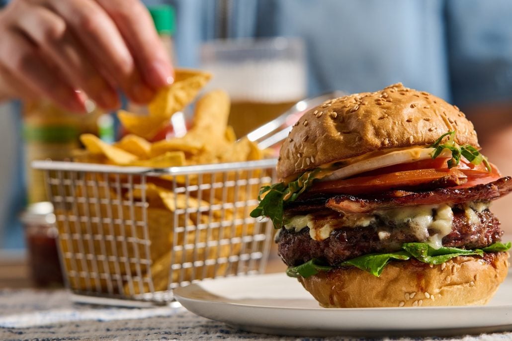 A cheeseburger with lettuce, tomato, onion, and bacon on a sesame seed bun is on a plate—just the kind of mouthwatering fare you might enjoy at the Beach Club on Holland America’s private island, Half Moon Cay. In the background, a metal basket holds fries as a hand reaches for them, while a drink sits partially visible behind the basket.