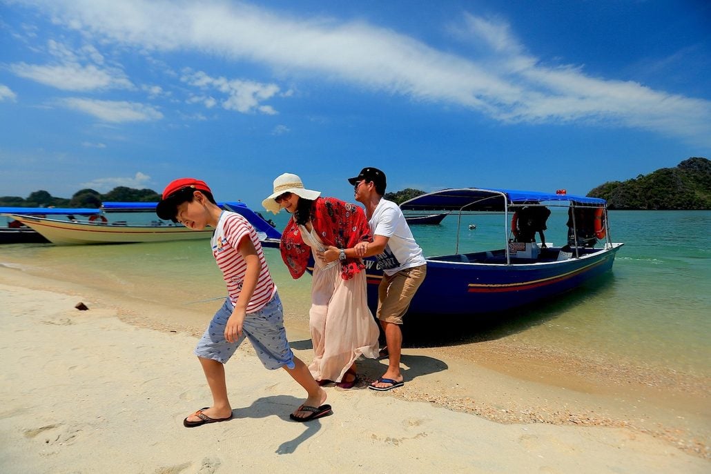 Three people are stepping off a blue boat onto a sandy beach in Langkawi; the sky is mostly clear with small clouds, and the water is calm and greenish-blue. One person assists another while a child walks ahead—a perfect scene for first-time visitors following any travel guide to this beautiful island.