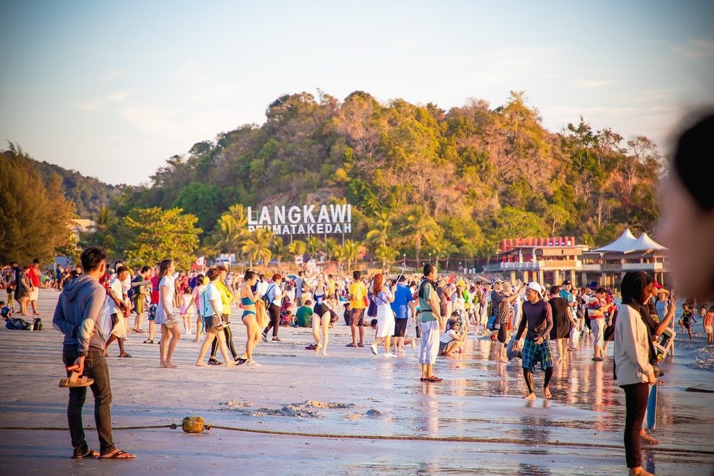A crowded beach scene at Langkawi, Pantai Cenang, Malaysia, with people walking on wet sand, trees and a hill in the background, and the “Langkawi” sign visible on the hillside—an idyllic spot often recommended in any travel guide for first-time visitors to Langkawi.