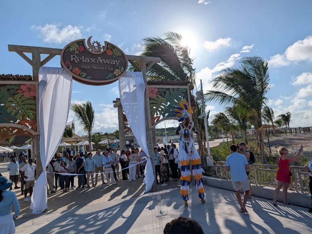 A crowd gathers at an outdoor event under a sign reading “RelaxAway Half Moon Cay.” A performer on stilts in a colorful costume stands in front, entertaining guests as they explore the free vs extra activities available. Palm trees and blue sky with clouds are in the background.