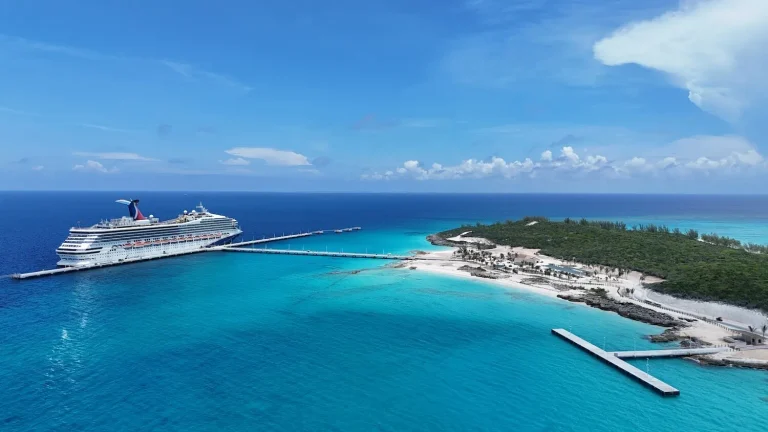 A large cruise ship is docked at a pier near Half Moon Cay, a small, sandy island with clear turquoise water and lush green vegetation under a bright blue sky with scattered clouds—a perfect scene for those seeking to RelaxAway.