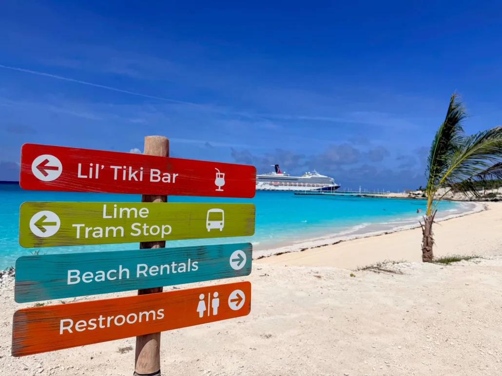Colorful sign on the sandy beach at Half Moon Cay points to a tiki bar, tram stop, beach rentals, and restrooms. Turquoise ocean, a cruise ship, palm tree, and blue sky are visible in the background—capturing that perfect RelaxAway vibe.