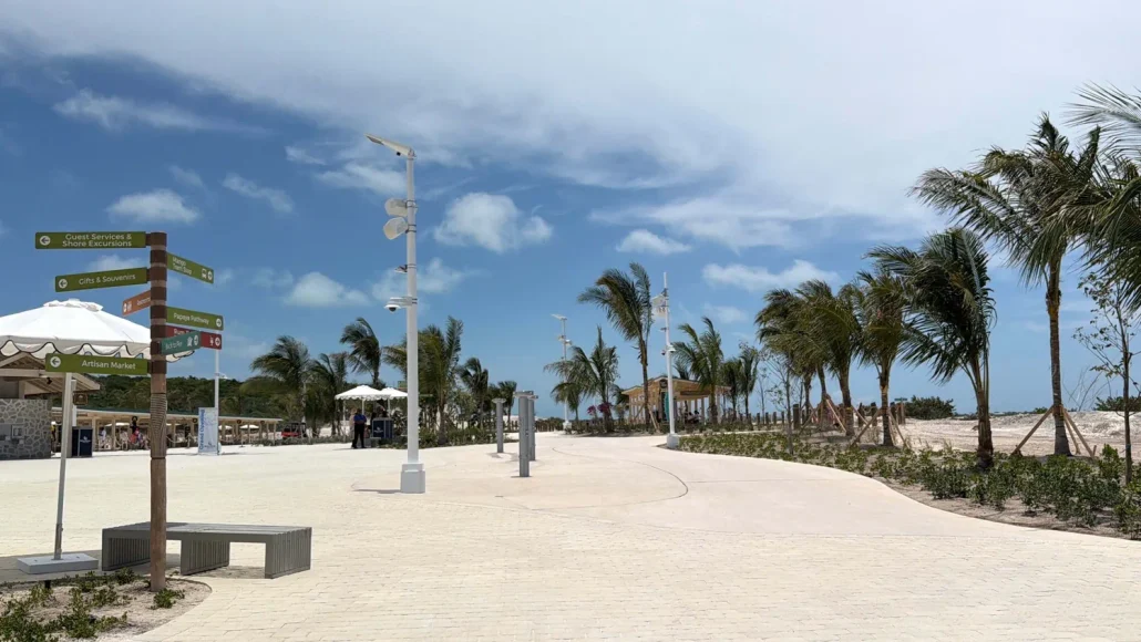A paved walkway lined with palm trees curves along a beach area under a partly cloudy sky on RelaxAway’s scenic Half Moon Cay. Directional signs, benches, and lamp posts are visible along the path, offering both free vs extra amenities for visitors’ convenience. A few people are in the distance.