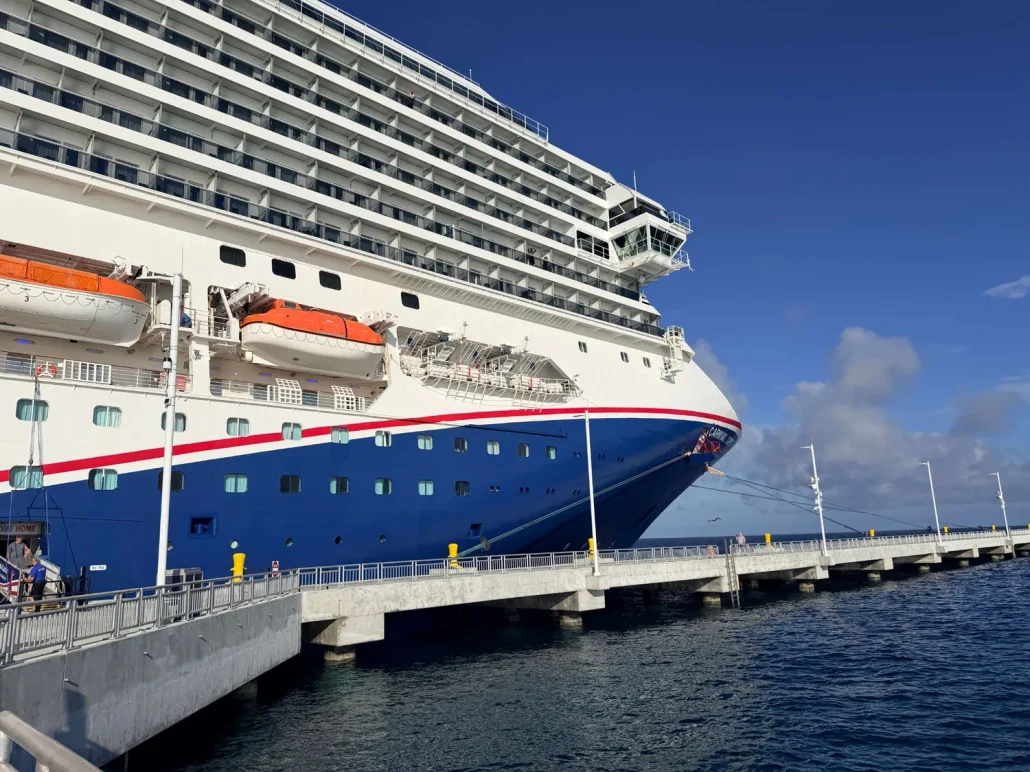 A large cruise ship docked at a pier under a clear blue sky, offering stunning views of Half Moon Cay. The ship’s white and blue exterior features orange lifeboats, while the pier extends over calm, dark blue water. Guests can explore RelaxAway amenities onboard, with clear options for free vs extra services during their stay.