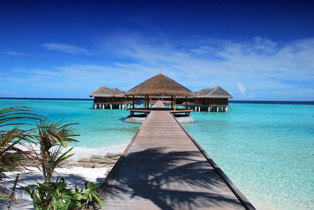 A wooden pier leads to overwater thatched-roof huts at an all-inclusive resort, surrounded by clear turquoise water and a sandy beach with palm trees under a sunny blue sky.