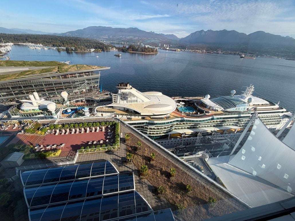 A large cruise ship docked at a port with pools and deck chairs visible, viewed from above. The background shows water, buildings, forested land, and mountains under a clear sky.