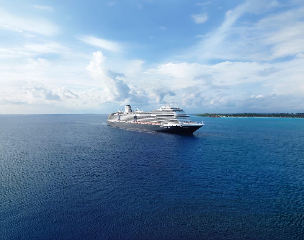 A large cruise ship sails on calm blue ocean waters under a partly cloudy sky, with a stretch of green coastline visible in the background—just beyond lies Half Moon Cay, where guests can enjoy the island’s famous free BBQ pavilion and unwind at the exclusive beach club.