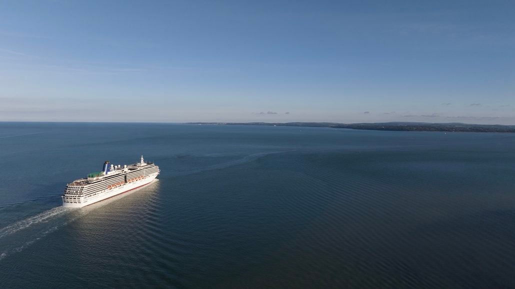A large Holland America cruise ship sailing through calm blue water toward a distant coastline.